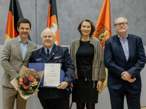 Vier Personen, darunter ein Herr mit Urkunde und Blumenstrauß