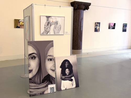 In einem Saal steht ein kurzes Wandelement, daran hängt ein gezeichnetes Bild mit zwei alten Händen, die ein Pflaster auf ein junges Knie kleben. Davor stehen zwei Bilder angelehnt, eines mit einer Frau und einem Mädchen in angeschnittenen Gesichtshälften, beide tragen Kopftuch. Daneben auf dem Bild eine dunkelhäutige Frau umfasst eine jüngere Frau von hinten um die Schultern, beide lächeln in die Kamera.