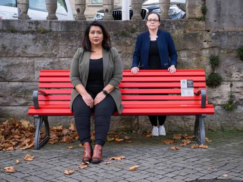 Eine Frau sitzt auf einer roten Bank, eine zweite Frau steht dahinter.