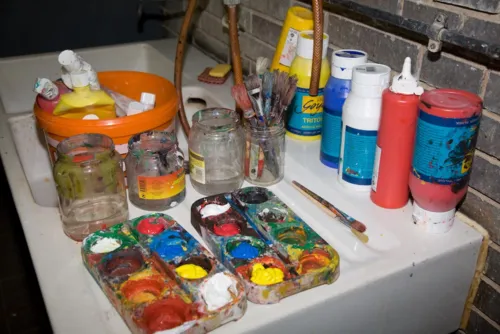 Farbpaletten mit Acrylfarbe drin, Pinsel und Gläser auf einem Waschbecken
