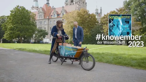 Ein Mann mit Perücke in Kleidung aus der Barockzeit auf einem Lastenrad und ein Mann im Anzug vor historischem Gebäude
