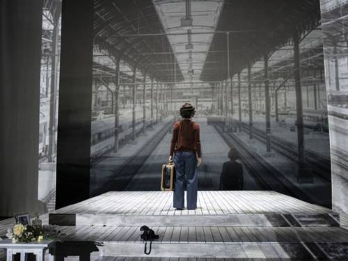 Eine Schauspielerin steht mit einem Koffer in der Hand auf einer Bühne, auf deren Wände Bahnsteige eines Bahnhofs projiziert sind.