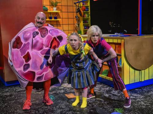 In einer bunten Kinderküche stehen auf einer Bühne zwei Schauspielerinnen als Mutter und Kind sowie ein Mann in einem Wurstscheibenkostüm.