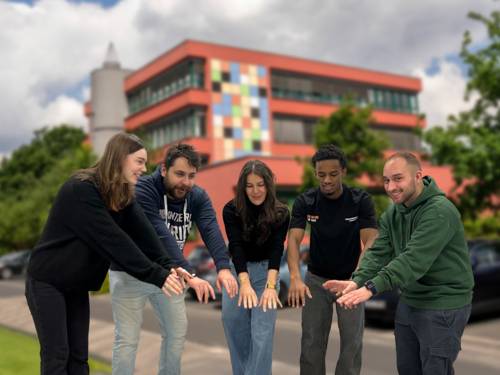 Fünd junge Menschen strecken vor dem Gebäude des Diakonie-Kolleg Hannover die Hände in Hüfthöhe voreinander aus und bilden dabei einen Viertelkreis.