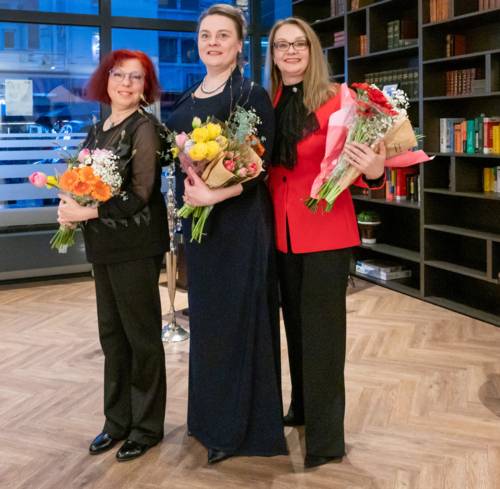 Drei Frauen in Abendgarderobe stehen vor einem Regal und halten Blumen in den Armen.