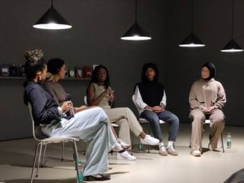 Fünf junge Menschen, vier Frauen und eine nichtbinäre Person, sitzen in einem Halbkreis auf weißen Stühlen. Die Personen sind einander zugewandt, die mittlere spricht in ein Mikrofon. Alle sind Persons of Color, einige schwarz-, einige braun-gelesen. Sie sitzen vor einer dunkelgrauen Wand mit einigen Büchern auf einem einzelnen Regal.
