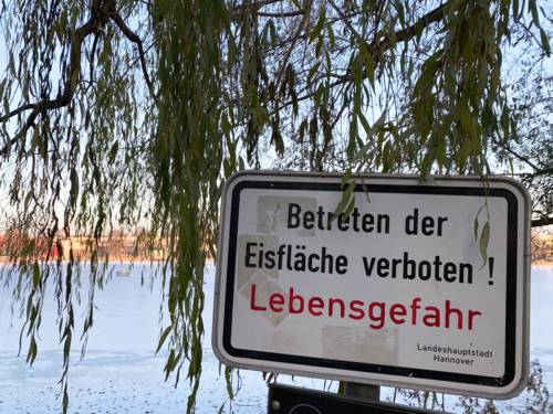 Ein weißes Schild mit der Aufschrift "Betreten der Eisfläche verboten! Lebensgefahr", im Hintergrund der zugefrorene Maschsee.