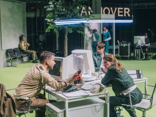 Eine Schauspielerin und zwei Schauspieler in Polizeiuniformen an Schreibtischen auf einer B&uuml;hne unterhalten sich.
