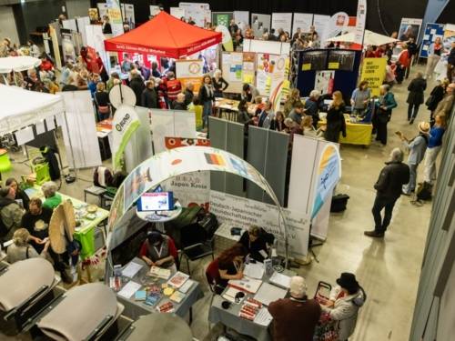 Blick von oben in einen großen Raum, in den sich viele Menschen zwischen Ständen tummeln. Rechts stehen zwei Musiker*innen.