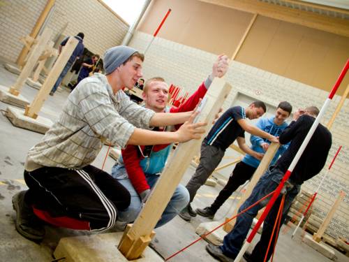 Zwei Berufsschüler messen einen senkrecht aufgekeilten Holzbalken mit einem Gliedermaßstab, sie sind in der Praxishalle ihrer Berufsschule.