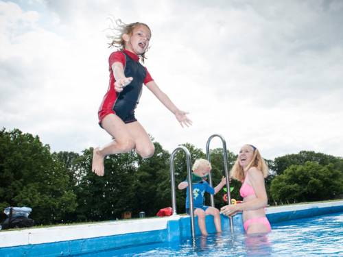 Ein Kind springt ins Wasser, ein anderes Kind und eine Frau sind im Wasser und am Beckenrand neben einer Leiter