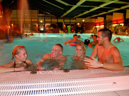 Zwei junge Frauen und zwei junge Männer am Rand eines Schwimmbeckens. Alle vier lächeln und im Wasser sind weitere Badegäste und zwei Wasserfontänen zu sehen.