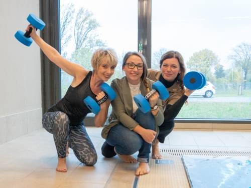 Drei Damen, teils in sportlicher Kleidung, sitzen am Rand eines Schwimmbeckens und halten blaue Hanteln in den Händen.