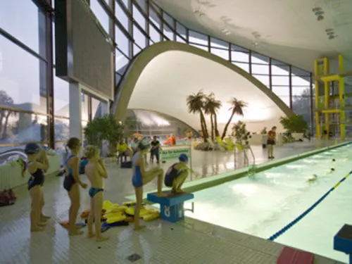 Kinder stehen wartend in einer Reihe, um ins Schwimmerbecken zu springen, im Hintergrund einige Palmen vor einer großen Fensterfront