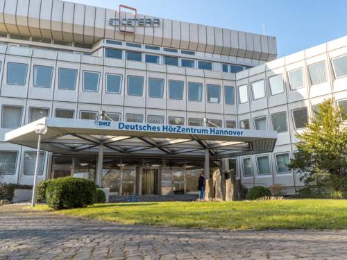 Klinikgebäude mit weißer Fassade und trapezförmigen Fenstern. Am Dach des Eingangs steht "DHZ Deutsches HörZentrum Hannover".