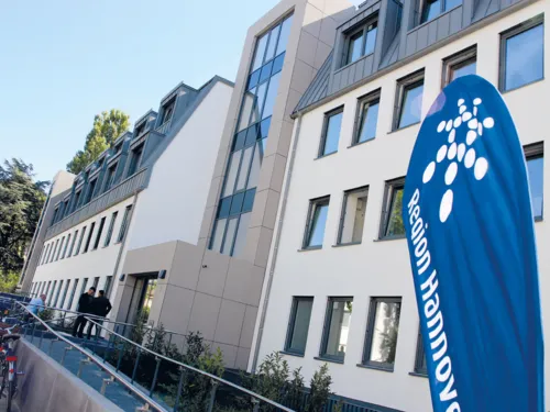 Ein modernes Gebäude mit einer Rampe für einen barrierefreien Zugang. Im Eingangsbereich stehen zwei Personen und eine weitere Person schließt ihr Fahrrad an. Vorne rechts im Bild ist ein blauer Aufsteller mit der Aufschrift "Region Hannover" zu sehen (das Wort Hannover ist zum Teil abgeschnitten).