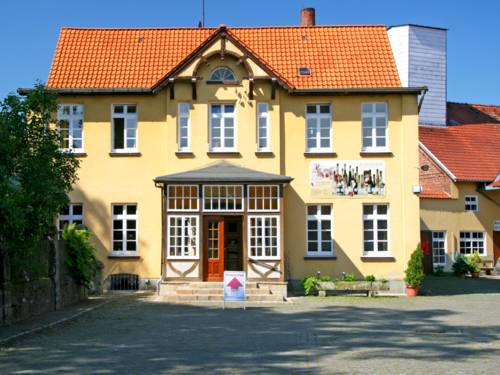 Zweigeschossiges Gebäude mit rotem Ziegeldach und gelber Fassade. Der Eingang führt durch einen Erker. An der linken Gebäudeseite ist ein Banner befestigt, auf dem verschiedene Glasflaschen zu sehen sind.