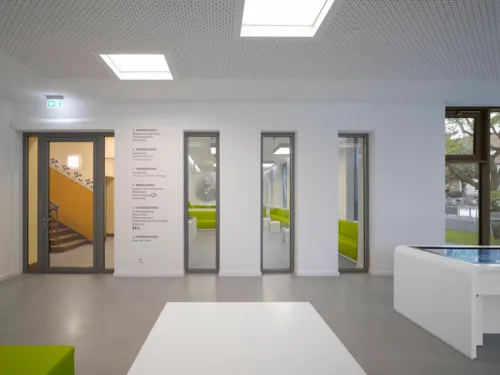 Ein Foyer mit einem Medientisch. Rechts neben der Tür, die in ein Treppenhaus und zu weiteren Räumlichkeiten führt, ist an die Wand geschrieben, was in den einzelnen Stockwerken zu finden ist.
