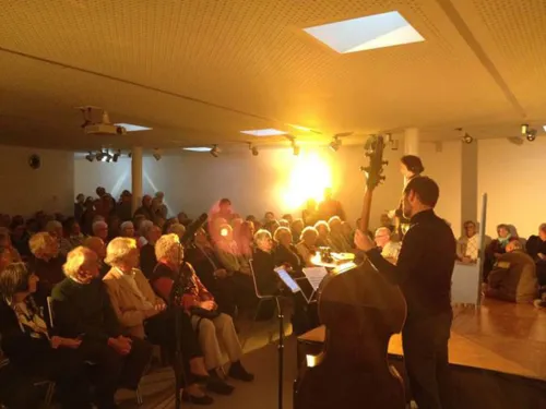 Ein kleiner Veranstaltungsraum, indem alle Sitzplätze belegt sind. Auf der Bühne stehen Musiker.