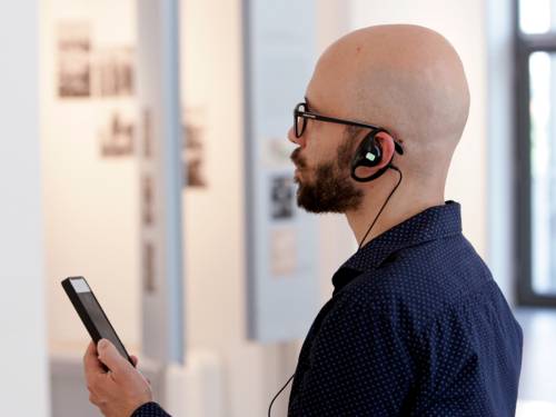 Ein Mann mit Bart und Brille steht mit einem Smartphone in der Hand und Kopfhörern im Ohr in einem hellen Raum