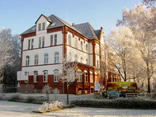 Gebäude über dreieinhalb Vollgeschosse. Die Fassade besteht teils aus roten Backsteinen, teils aus weiß verputzten Flächen. Die umstehenden Bäume sind von Eis überzogen und strahlen im Sonnenschein winterlich weiß.