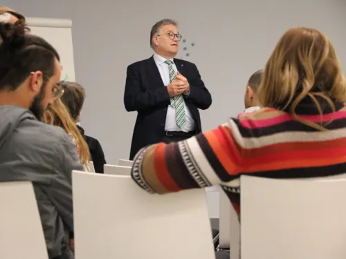 Ein Mann im Anzug steht vor sitzenden Menschen und redet.