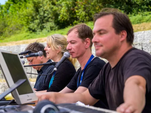 Vier Personen in schwarzen T-Shirts sitzen im Freien und sprechen in Mikrofone vor ihnen.