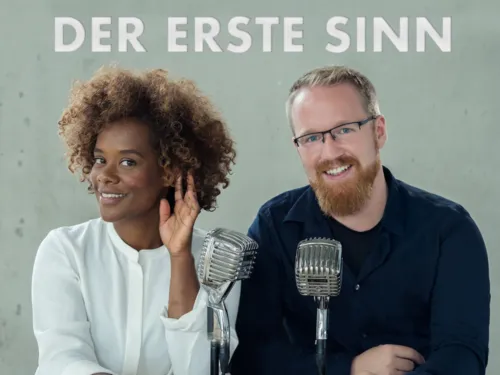 Eine Frau mit Locken und ein Mann mit Bart und zwei Mikrofone im Vordergrund.