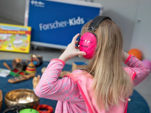 Ein Mädchen trägt Kopfhörer, im Hintergrund ist die Entdeckerkiste der Forscherkids zum Thema Klänge und Geräusche zu sehen, Instrumente und andere klingende Gegenstände oder Material für Hör-Experimente liegt daneben.