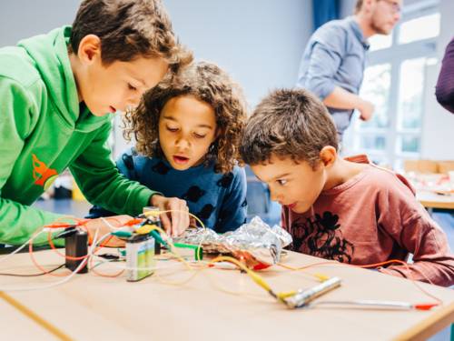 Drei Kinder experimentieren an einem Tisch mit Drähten, Batterien, Metallen und weiteren Utensilien.