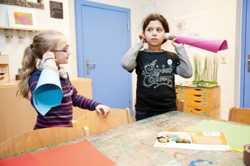 Zwei Kinder, die sich zusammengerolltes Papier an die Ohren halten um besser zu hören