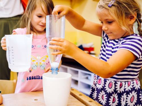 Zwei Mädchen experimentieren mit Wasser, einer aufgeschnittenen Plastikflasche und weiteren Gefäßen.