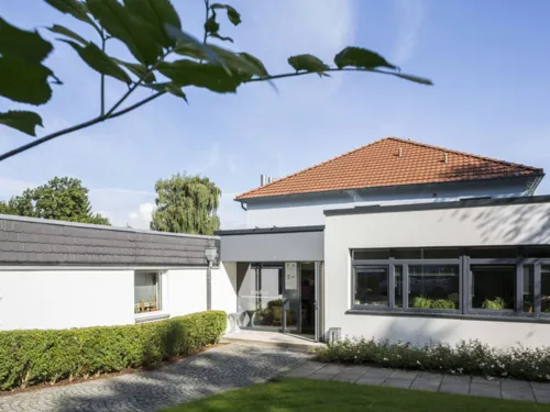 Eingangsbereich und Gebäudeteile mit Flachdach, die weiß verputzt sind. Dahinter ist ein weiteres Gebäude zu sehen, das ein Giebeldach mit roten Dachziegeln.