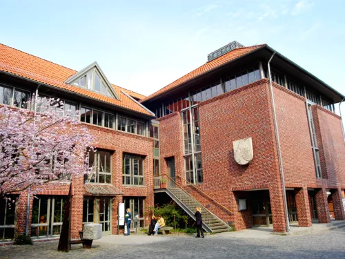 Großer Backsteinbau mit vielen Fenstern. Der blühende Kirschbaum lässt vermuten, dass das Bild im Frühjahr aufgenommen wurde.