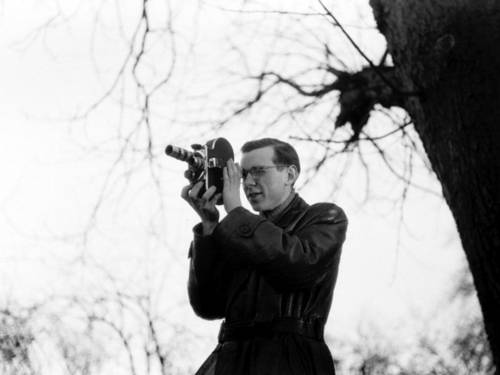 Ein Mann im Ledermantel und mit Brille steht draußen neben einem Baum und fotografiert mit einer Kamera.