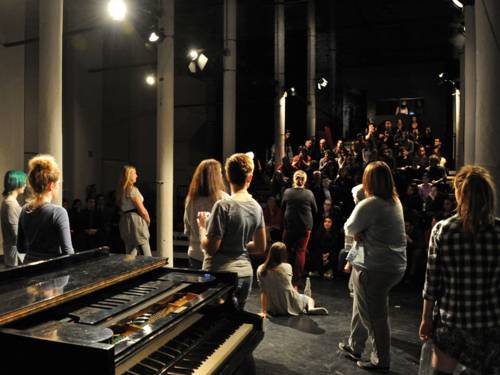 Ein Klavier und mehrere Mädchen auf einer Theaterbühne. Der Blick geht von der Bühne in Richtung Publikum - die Schauspielerinnen sind von hinten und die Zuschauer von vorne zu sehen.