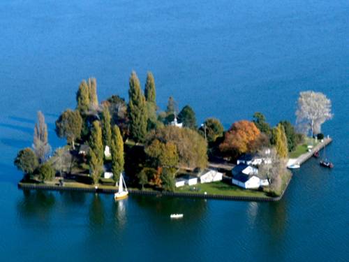 Luftaufnahme: Nahezu quadratische, flache Insel mit Bäumen und Gebäuden. Am Ufer ankern zwei Segelbote.