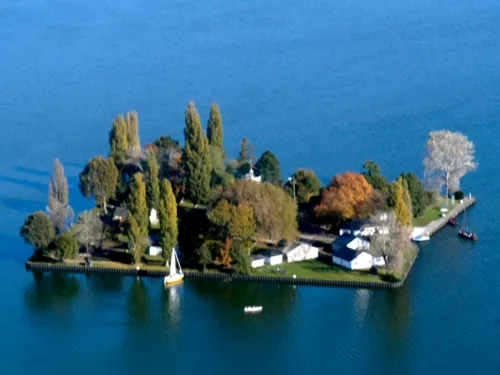 Luftaufnahme: Nahezu quadratische, flache Insel mit Bäumen und Gebäuden. Am Ufer ankern zwei Segelbote.