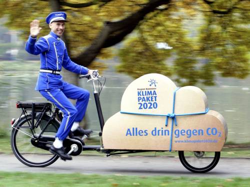 Ein Mann trägt eine Dienstbotenuniform und fährt auf einem Lastenfahrrad. Auf der Ladefläche des Fahrrads ist eine braune Wolke in Paketform. Darauf steht "Region Hannover. Klimapaket 2020. Alles drin gegen CO2 – www.klimapaket2020.de
