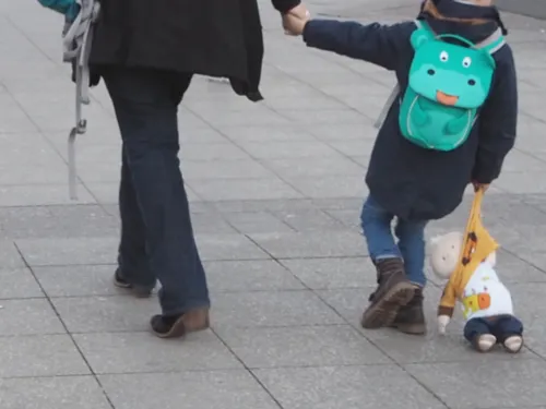 Ein Kind geht neben einem Erwachsenen. Mit der linken Hand hält es die rechte Hand des Erwachsenen, mit der rechten Hand zieht es eine Stoffpuppe hinter sich her, die Puppe schleift auf dem Boden.