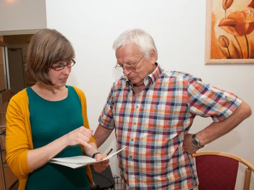 Eine Frau und ein Mann stehen beieinander, sie zeigt ihm etwas auf einem Blatt Papier, das sie vor sich hält.