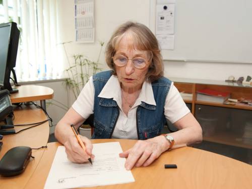 Eine ältere Frau sitzt an einem Tisch und füllt mit einem Stift ein Formular aus.