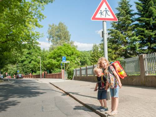 Zwei Kinder stehen am Straßenrand vor dem Verkehrsschild "Kinder" und schauen, ob die Straße, die sie offensichtlich queren wollen, frei ist
