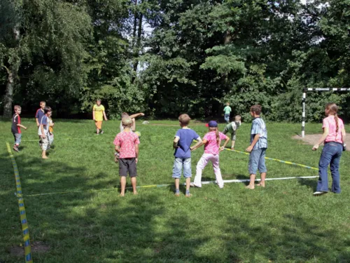 Auf einer Rasenfläche ist mit blau-gelbem Flatterband ein Spielfeld abgesteckt. Darauf spielen Kinder Völkerball: Die Kinder der gegnerischen Mannschaft werden mit einem Ball abgeworfen.