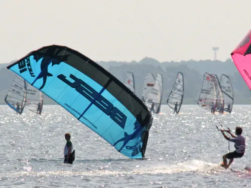 Eine Person steht auf einem Surfbrett und lässt sich von einem Lenkdrachen über das Wasser ziehen. Links ist ein blauer Lenkdrachen eines weiteren Kitesurfers zu erkennen, im Hintergrund sind Wassersportler als Windsurfer unterwegs.