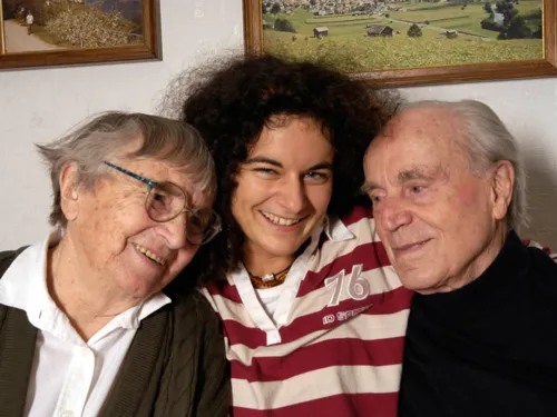 Eine junge Frau, die rechts im Arm eine Seniorin hält und links im Arm einen Senior. Hinten an der Wand sind Teilstücke zweier Gemälde zu sehen.