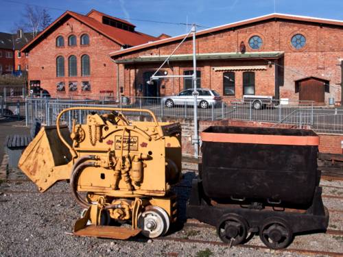 Auf dem Gelände des Besucherbergwerkes Barsinghausen steht ein kleines, sehr kompaktes Schienenfahrzeug für den Einsatz unter Tage. Hinter das gelbe Zugfahrzeug ist eine schwarze Güterlore gekoppelt.