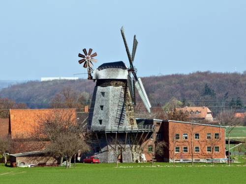Im Hintergrund der Windmühle sind landwirtschaftliche Gebäude und hügelige Landschaft zu erkennen.