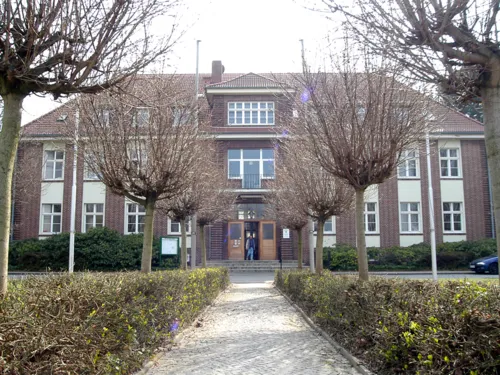 Fußweg gesäumt von einer Hecke und Bäumen auf ein dreistöckiges Gebäude zu