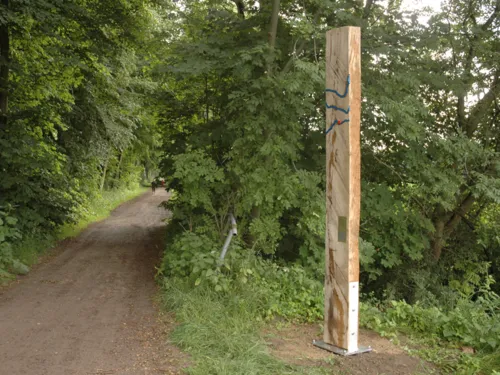 Senkrecht stehender Holzbalken mit blauen Metallwellen, die ein größeres Gewässer symbolisieren